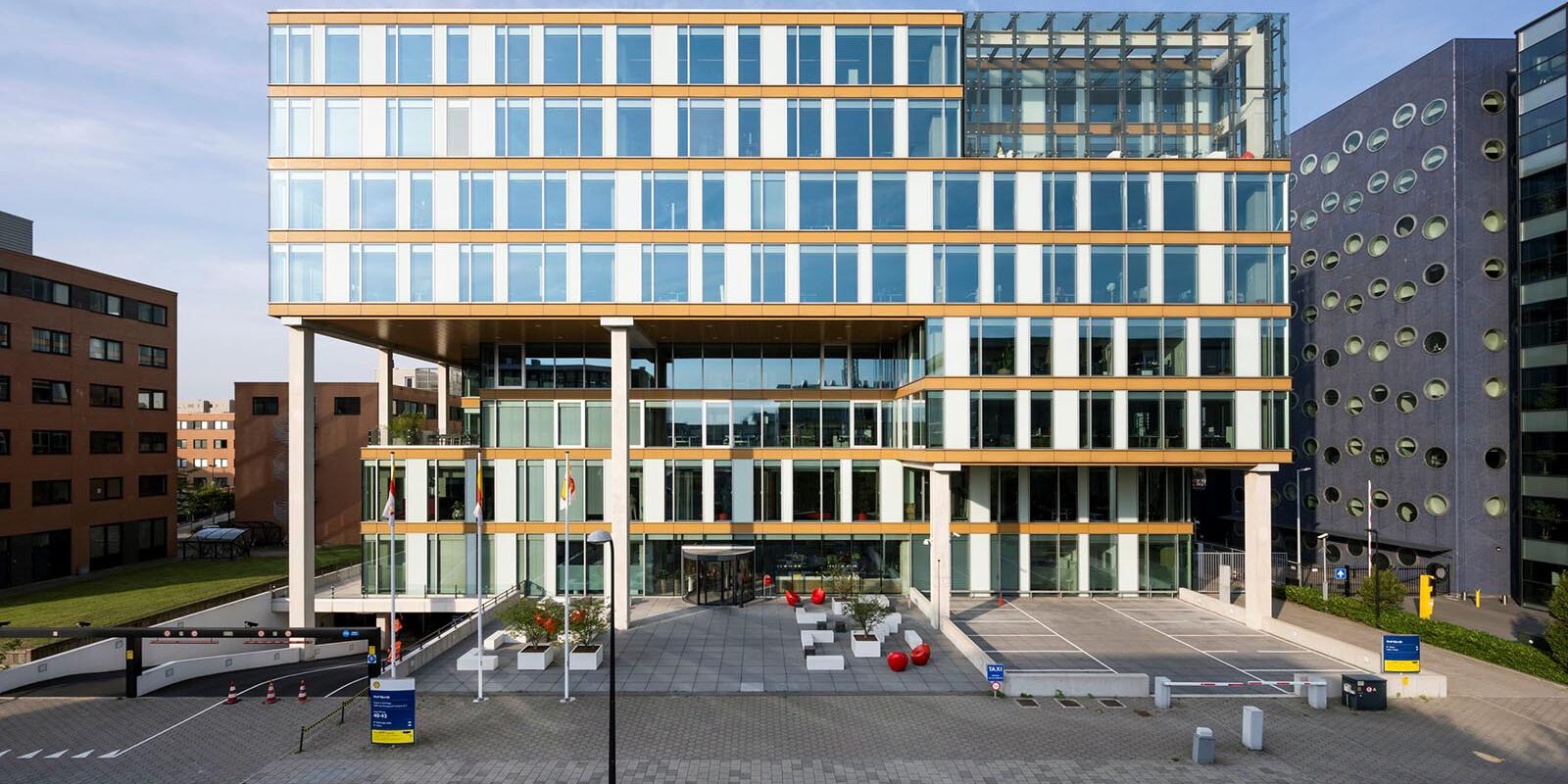 Modern multi-story office building with glass facade, outdoor seating area with red chairs, and parking entrances on a sunny day.