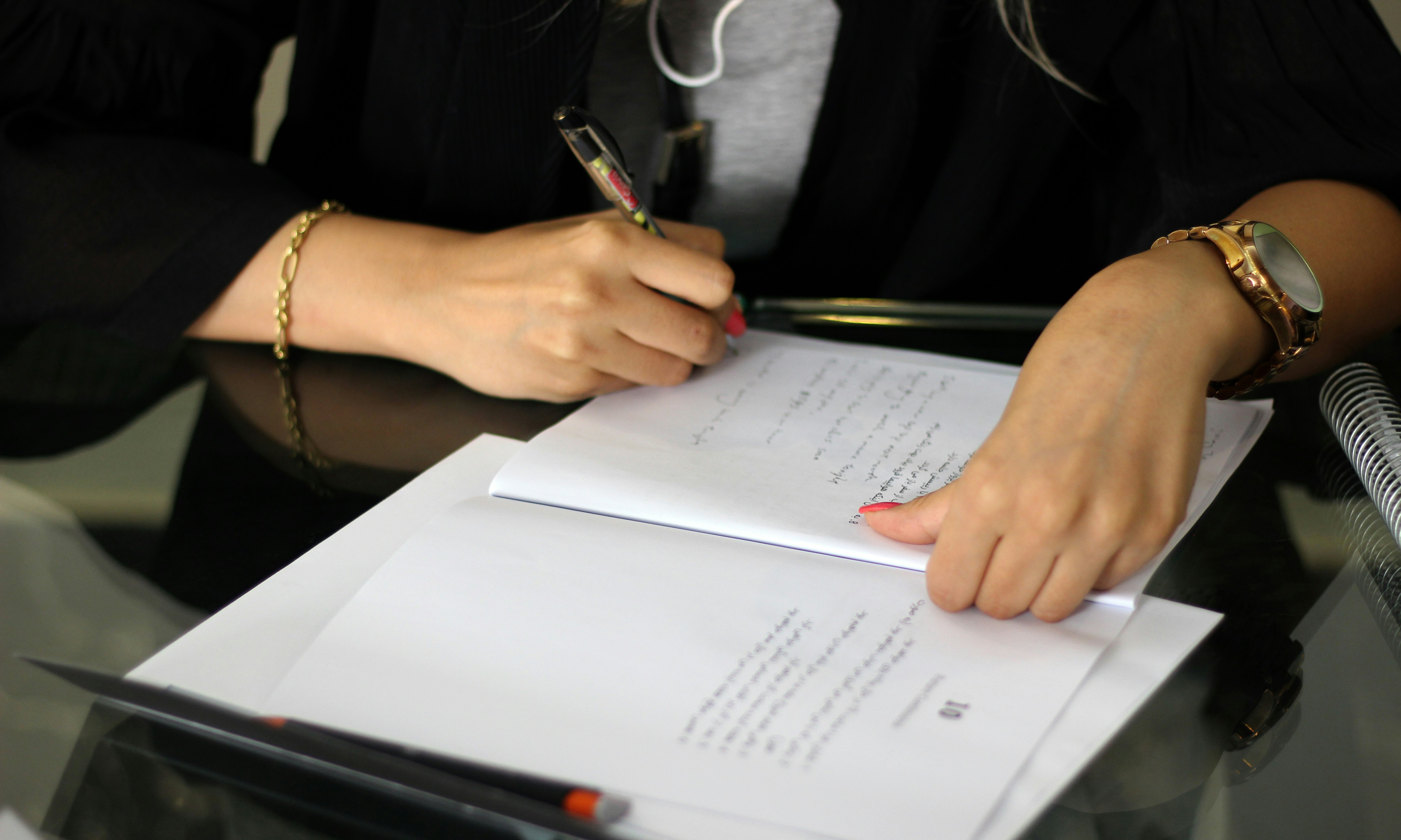 Person writing notes with a pen on papers laid on a glass table.