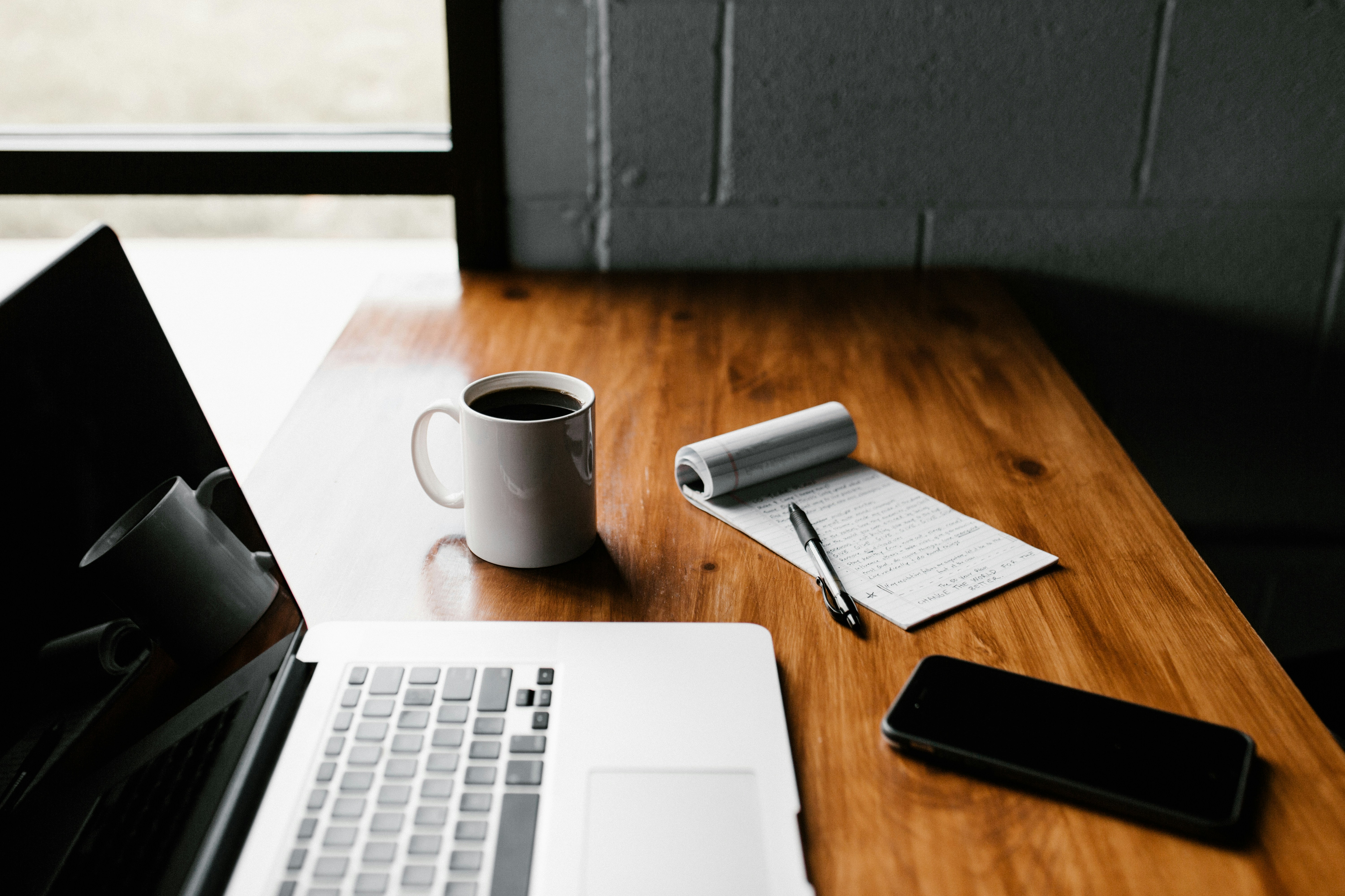 White laptop, coffee mug, notepad with pen, and smartphone on a wooden desk near a window.