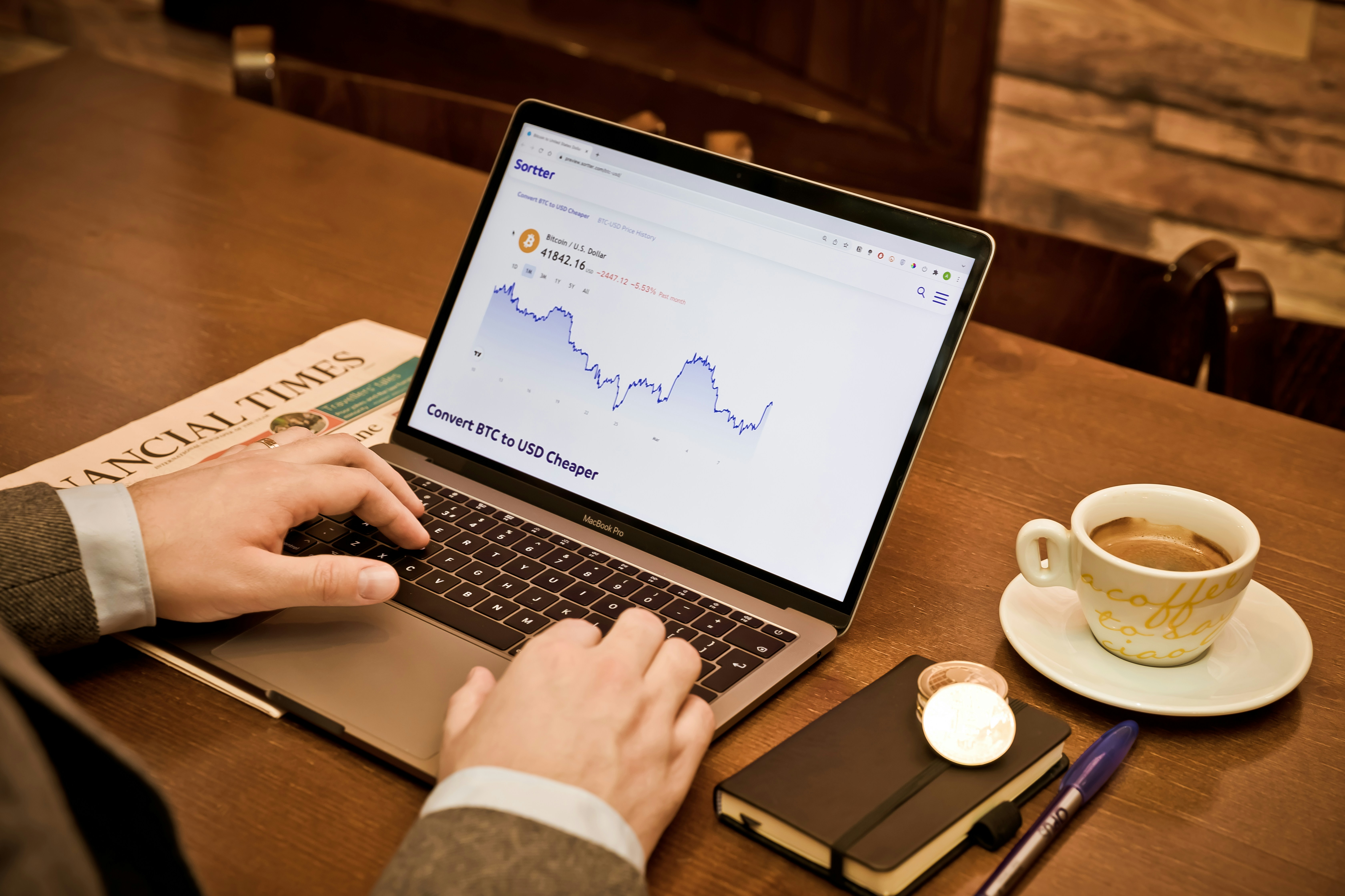 Person using a MacBook Pro showing Bitcoin to USD exchange rate graph, with a Financial Times newspaper, a notebook, a pen, and a coffee cup on a wooden table.