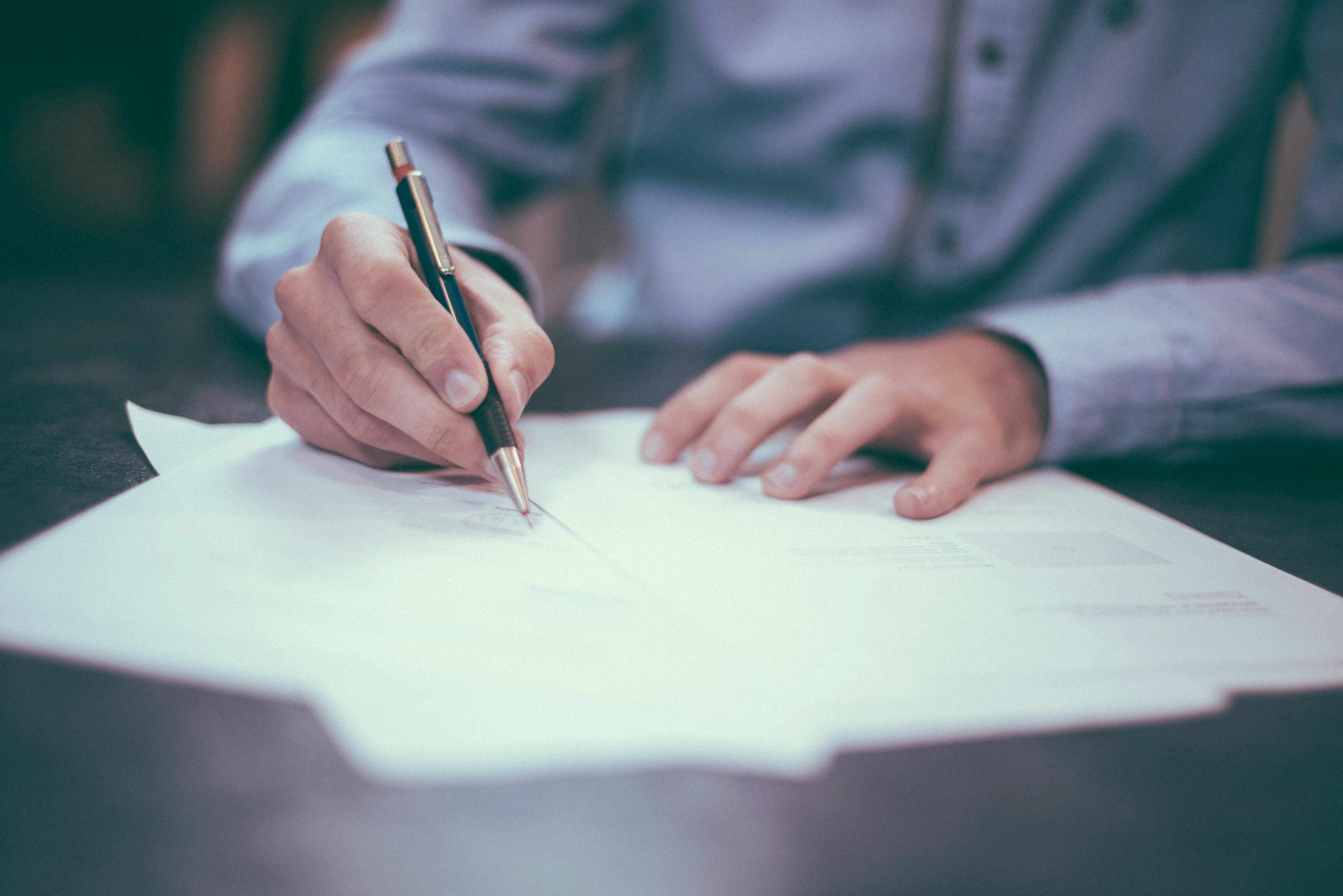 Person in a blue shirt writing on a stack of white papers with a black pen.