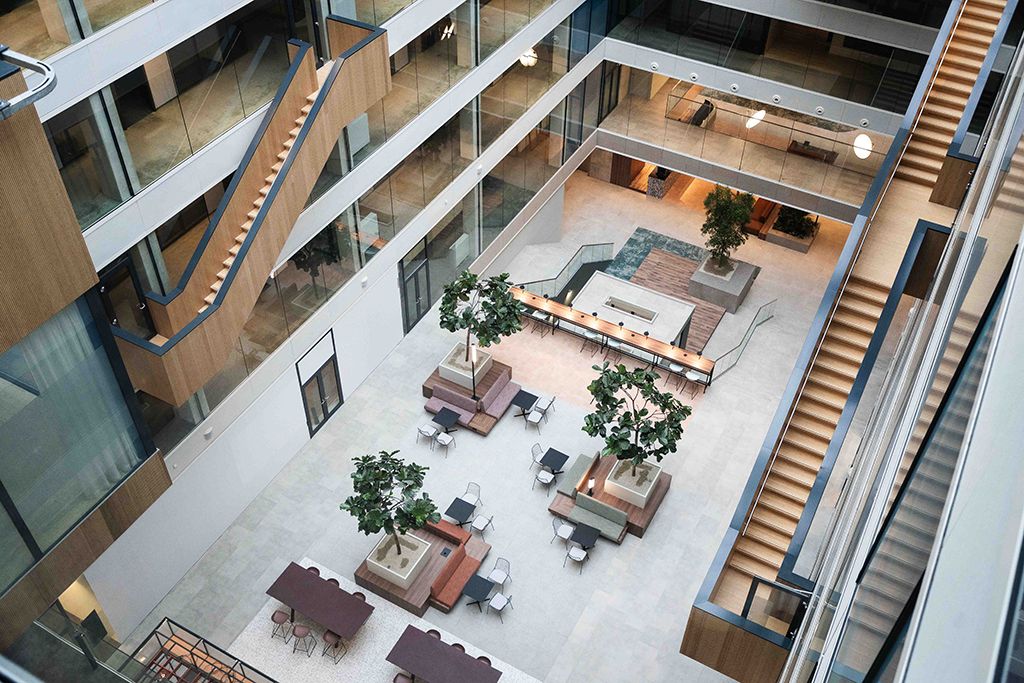 Modern multi-story office atrium with indoor trees, seating areas, long wooden tables with stools, and staircases with glass railings.